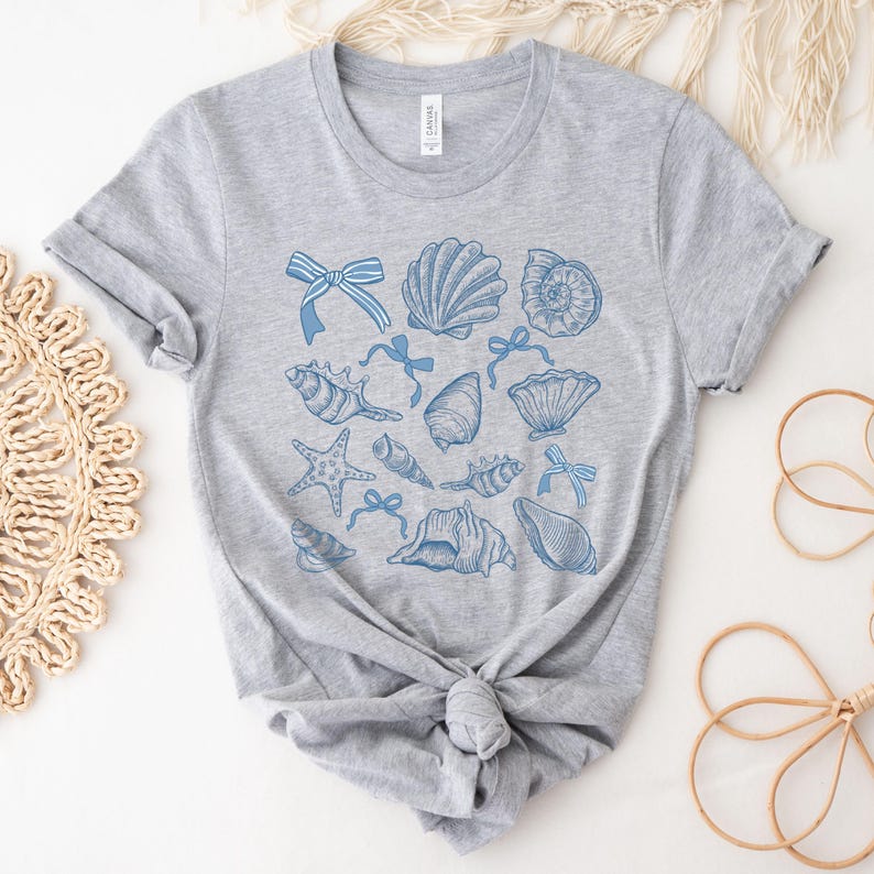 Pu&ograve; includere: Una maglietta grigia con un design a conchiglie blu e bianche. Il design presenta varie conchiglie, stelle marine e fiocchi.