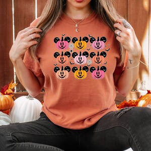 May include: A burnt orange t-shirt with a graphic of nine orange, pink, and peach Mickey Mouse pumpkins with black outlines. The pumpkins have black stems and black Mickey Mouse ears. The graphic is surrounded by black bats.