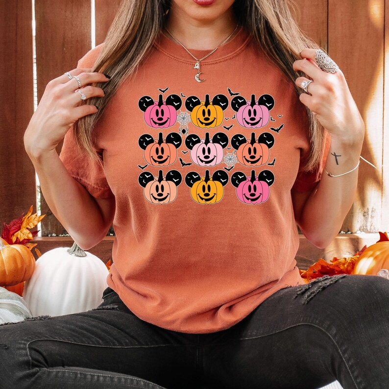 May include: A burnt orange t-shirt with a graphic of nine orange, pink, and peach Mickey Mouse pumpkins with black outlines. The pumpkins have black stems and black Mickey Mouse ears. The graphic is surrounded by black bats.