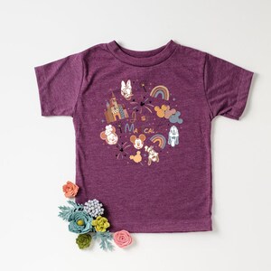 以下が含まれることがあります： 紫色の半袖Tシャツに、漫画の城、ミッキーマウスのキャラクター、そして「It's Magical」というテキストのグラフィックデザインが施されています。