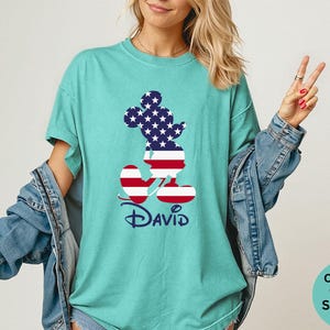 May include: Seafoam green t-shirt featuring a patriotic Mickey Mouse design. The silhouette is filled with the American flag pattern. The name "David" is printed below the character in a script font. The shirt is part of the Comfort Colors collection.