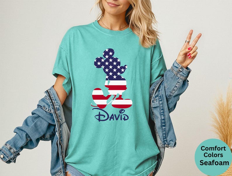 May include: Seafoam green t-shirt featuring a patriotic Mickey Mouse design. The silhouette is filled with the American flag pattern. The name "David" is printed below the character in a script font. The shirt is part of the Comfort Colors collection.