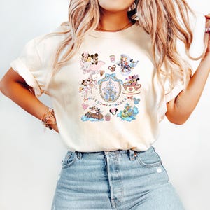 May include: Cream-colored t-shirt featuring a colorful graphic of Disney characters, including Mickey and Minnie Mouse, Donald Duck, and others. The design includes a castle, teacups, and the text "Best Day Ever".