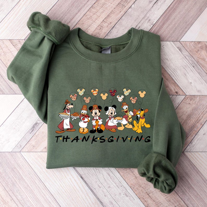K&ouml;nnte beinhalten: Gr&uuml;ner Pullover mit Thanksgiving-Motiv mit Mickey Mouse, Minnie Mouse, Donald Duck, Goofy und Pluto. Die Figuren halten Kuchen und sind von Mickey Mouse-f&ouml;rmigen Ballons umgeben. Der Text "THANKSGIVING" ist unter den Figuren gedruckt.