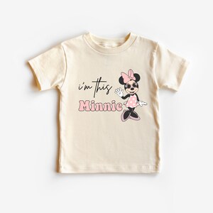 K&ouml;nnte beinhalten: Cremefarbenes T-Shirt f&uuml;r Kleinkinder mit einer Grafik von Minnie Mouse mit Sonnenbrille und einem rosa gepunkteten Kleid. Der Text "i'm this Minnie" ist in Schwarz und Rosa auf der Vorderseite aufgedruckt.