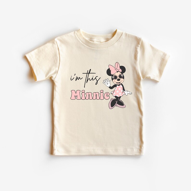 K&ouml;nnte beinhalten: Cremefarbenes T-Shirt f&uuml;r Kleinkinder mit einer Grafik von Minnie Mouse mit Sonnenbrille und einem rosa gepunkteten Kleid. Der Text "i'm this Minnie" ist in Schwarz und Rosa auf der Vorderseite aufgedruckt.