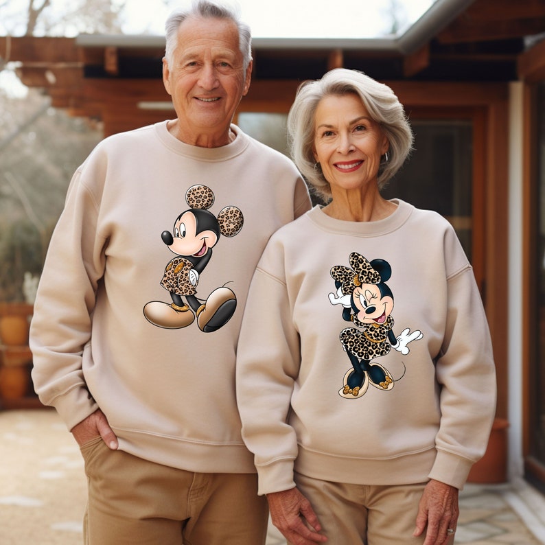 Op de afbeelding: Een stel draagt ​​passende beige sweatshirts met een cartoonafbeelding van Mickey Mouse en Minnie Mouse die kleding met luipaardprint dragen.