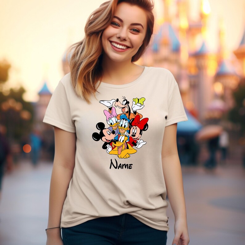 Op de afbeelding: Een beige T-shirt met een kleurrijke afbeelding van Mickey Mouse, Minnie Mouse, Donald Duck, Goofy, Pluto en Daisy Duck. De tekst "Name" staat onder de afbeelding.