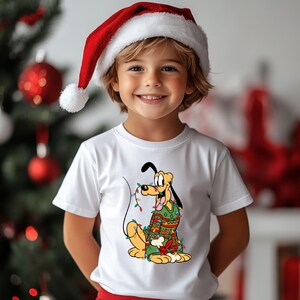 May include: A child wearing a white t-shirt featuring a cartoon Pluto character wrapped in Christmas lights and a green sweater. The child is also wearing a red and white Santa hat. A Christmas tree is visible in the background.