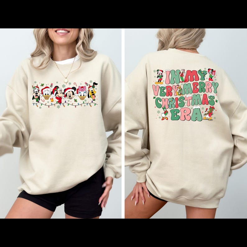 May include: A beige sweatshirt with a Christmas-themed design featuring Mickey Mouse and other Disney characters on the front. The back of the sweatshirt has the text "In My Very Merry Christmas Era" with a festive design.