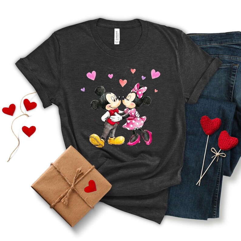 May include: Dark grey t-shirt with a watercolour illustration of Mickey and Minnie Mouse kissing, surrounded by pink hearts. The shirt is styled with blue jeans and a wrapped gift box with a red heart. Decorative red heart-shaped ornaments are also present.