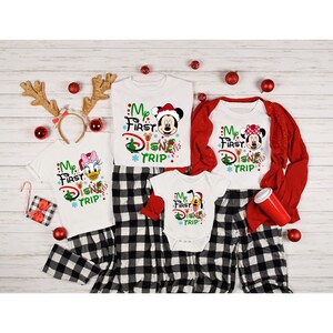 May include: A family of four wearing matching white shirts with a Christmas-themed "My First Disney Trip" design. The shirts feature different Disney characters, including Mickey Mouse, Minnie Mouse, Donald Duck, and Pluto. The family is wearing matching black and white plaid pants.
