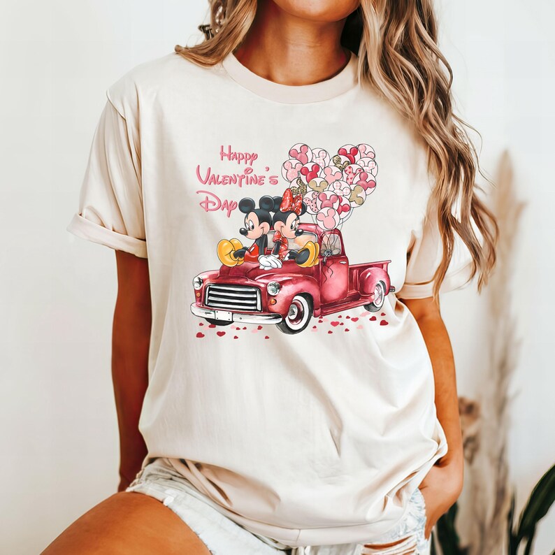 May include: A beige t-shirt with a graphic of Mickey Mouse and Minnie Mouse in a red pickup truck with pink heart balloons. The text "Happy Valentine's Day" is printed above the graphic.
