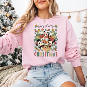 May include: A light pink sweatshirt featuring a Christmas graphic with Mickey Mouse, Minnie Mouse, Donald Duck, Goofy, and Pluto. The graphic includes Christmas trees and the text "Very Merry Christmas".