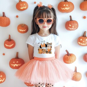 May include: A young person wearing orange sunglasses, a white t-shirt with a Halloween graphic, and a peach-colored tutu skirt. The background features carved pumpkins and orange accents, creating a festive Halloween theme.