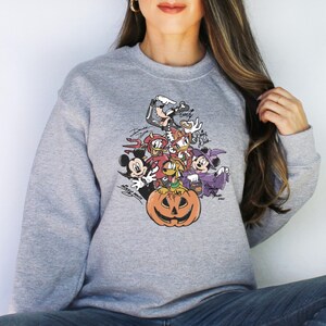 May include: A gray sweatshirt featuring a Halloween-themed graphic of Disney characters, including Mickey Mouse, Minnie Mouse, and Goofy, surrounding a carved pumpkin. The design includes character signatures.