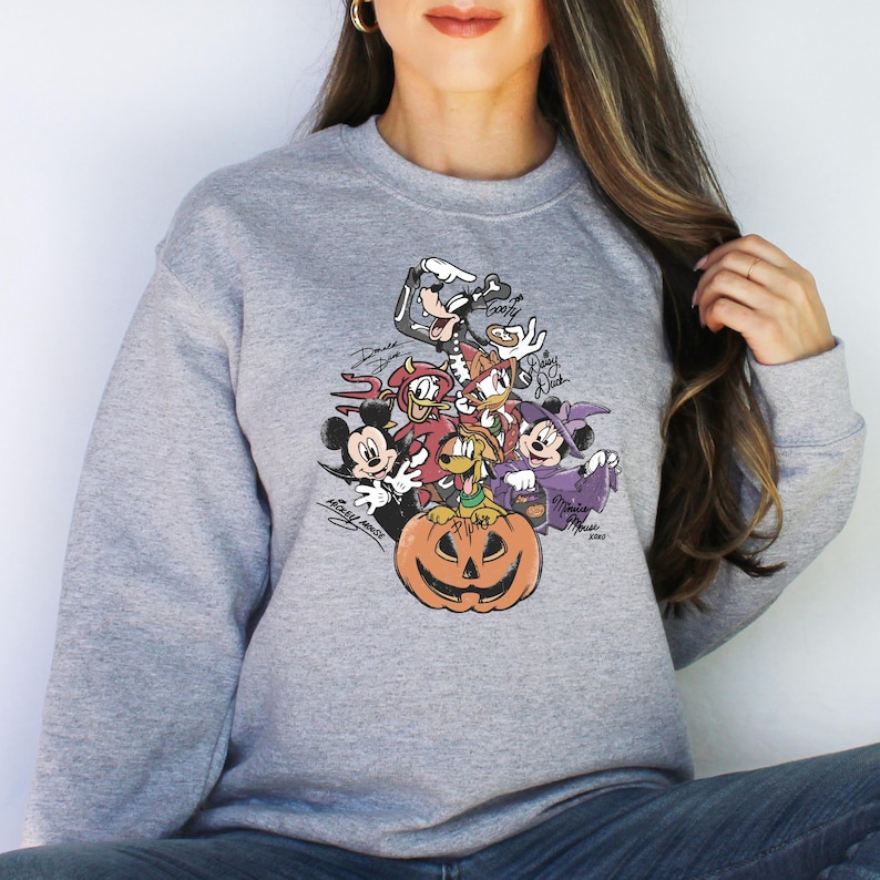 May include: A gray sweatshirt featuring a Halloween-themed graphic of Disney characters, including Mickey Mouse, Minnie Mouse, and Goofy, surrounding a carved pumpkin. The design includes character signatures.