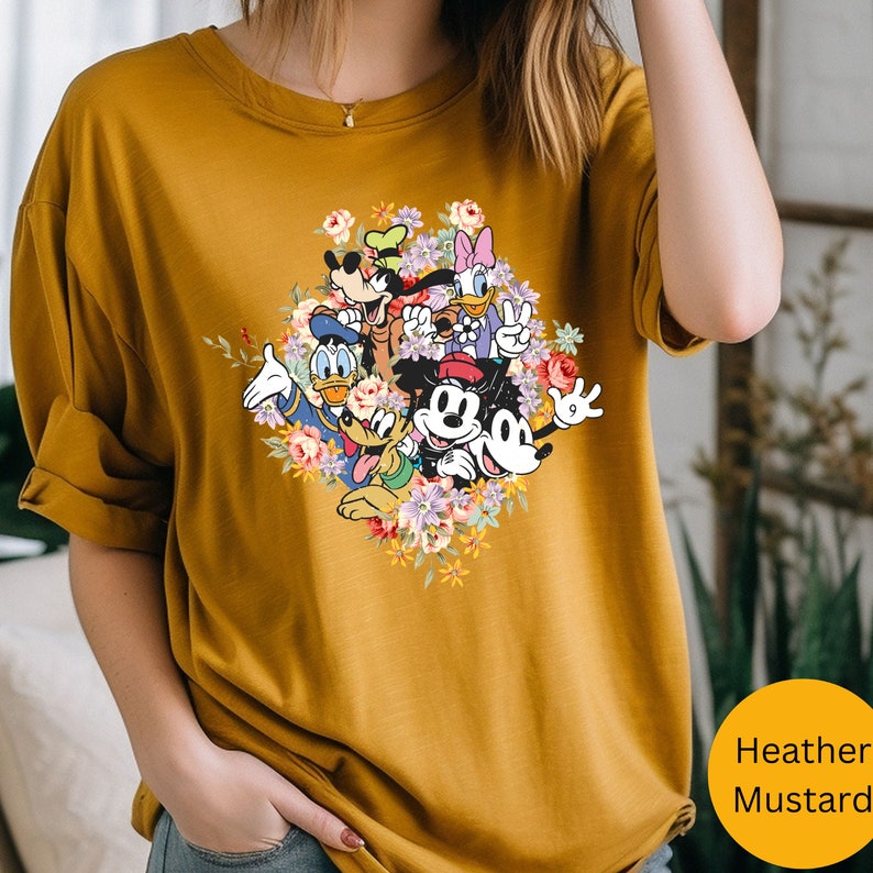 May include: A mustard yellow t-shirt with a colorful graphic of Mickey Mouse and his friends surrounded by flowers.