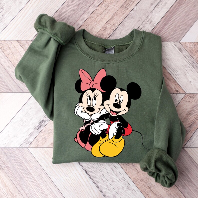 May include: A green sweatshirt with a graphic print of Mickey Mouse and Minnie Mouse. The characters are sitting together and smiling.