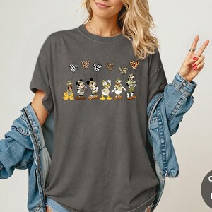 K&ouml;nnte beinhalten: Dunkelgraues T-Shirt mit Zeichentrickfiguren, die gemusterte Ballons halten. Zu den Figuren geh&ouml;ren ein Hund, eine Maus, eine Ente und andere. Das Shirt hat die Worte "Comfort Colors Pepper" auf einem runden Etikett.