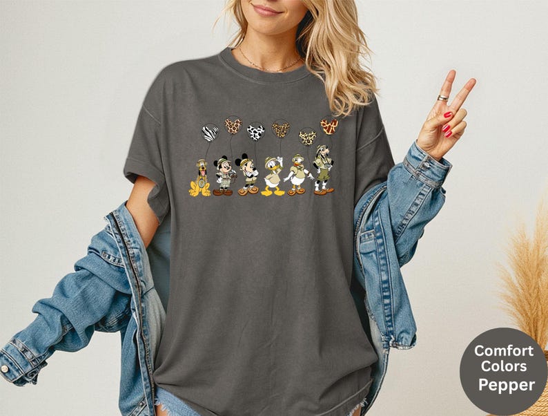 K&ouml;nnte beinhalten: Dunkelgraues T-Shirt mit Zeichentrickfiguren, die gemusterte Ballons halten. Zu den Figuren geh&ouml;ren ein Hund, eine Maus, eine Ente und andere. Das Shirt hat die Worte "Comfort Colors Pepper" auf einem runden Etikett.
