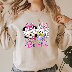 May include: A beige sweatshirt with a graphic print of Minnie Mouse and Daisy Duck. The graphic features the characters with their hands up in a peace sign. The text "Besties" is printed below the characters.