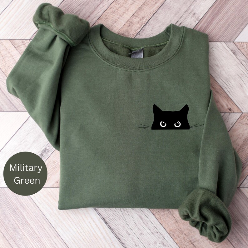 Pu&ograve; includere: Una felpa verde militare con un gatto nero che spunta dalla tasca. Il gatto ha baffi bianchi e grandi occhi neri.