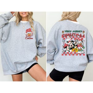 May include: A gray sweatshirt with a white collar. The front features a Donald Duck graphic with the word "Custom". The back displays a "Very Merry Christmas" design with Mickey Mouse, Goofy, Donald Duck, and other characters.