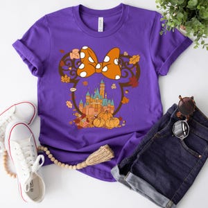 May include: Purple t-shirt with a graphic of a Mickey Mouse-inspired silhouette with a bow and a castle in the background. The castle is surrounded by fall leaves and pumpkins.