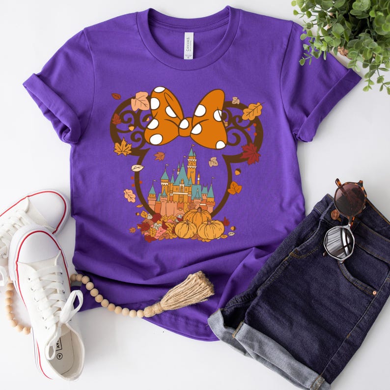 May include: Purple t-shirt with a graphic of a Mickey Mouse-inspired silhouette with a bow and a castle in the background. The castle is surrounded by fall leaves and pumpkins.