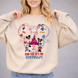 May include: Beige sweatshirt featuring a Mickey Mouse-shaped design with Disney characters, fireworks, and a castle. The design includes the text "4th of July" and "And Yes It's My Birthday."