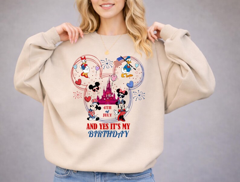 May include: Beige sweatshirt featuring a Mickey Mouse-shaped design with Disney characters, fireworks, and a castle. The design includes the text "4th of July" and "And Yes It's My Birthday."