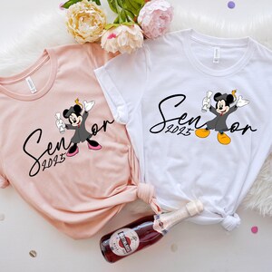 May include: Two white t-shirts with a pink t-shirt. The white shirts feature a black and white graphic of Mickey Mouse in a graduation cap and gown with the text "Senior 2023". The pink shirt has the same graphic but is in a smaller size.