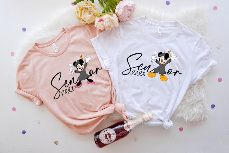 May include: Two white t-shirts with a pink t-shirt. The white shirts feature a black and white graphic of Mickey Mouse in a graduation cap and gown with the text "Senior 2023". The pink shirt has the same graphic but is in a smaller size.