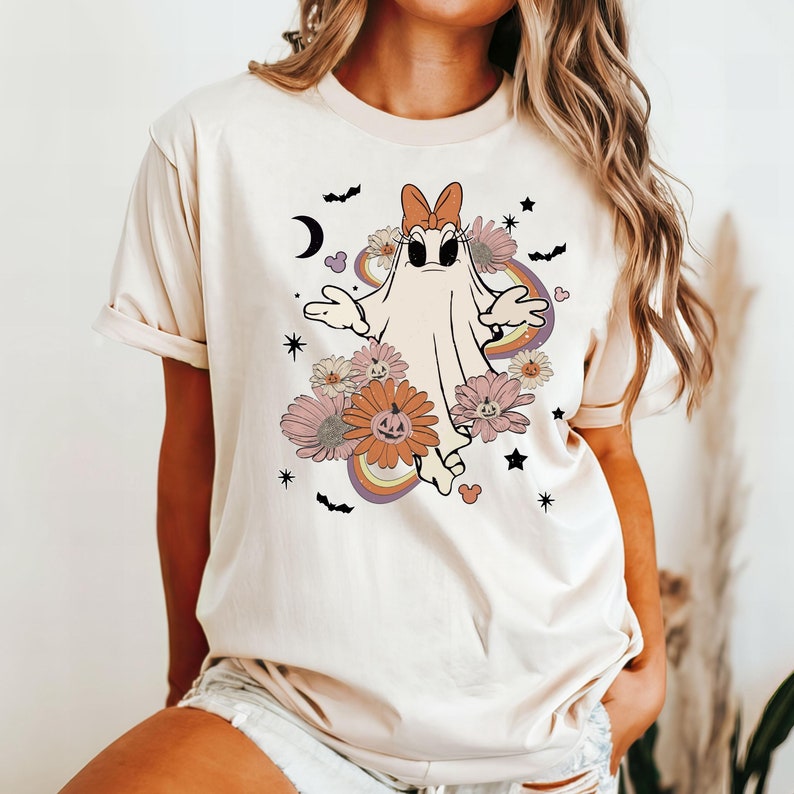 May include: A white t-shirt with a cartoon ghost wearing a pink bow and surrounded by flowers and pumpkins. The ghost has large eyes and a surprised expression. The design is in a retro style with rainbow accents.