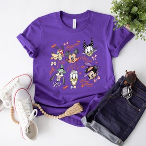 Puede incluir: Una camiseta morada con un dise&ntilde;o de Halloween con personajes de Disney, como Mickey y Minnie Mouse. La camiseta se combina con pantalones cortos de mezclilla y zapatillas blancas.