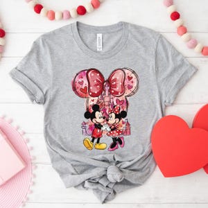 May include: A gray t-shirt featuring a graphic of Mickey and Minnie Mouse holding hands in front of a castle. The design includes a large bow with heart accents and a pink and red color scheme, perfect for Valentine's Day.