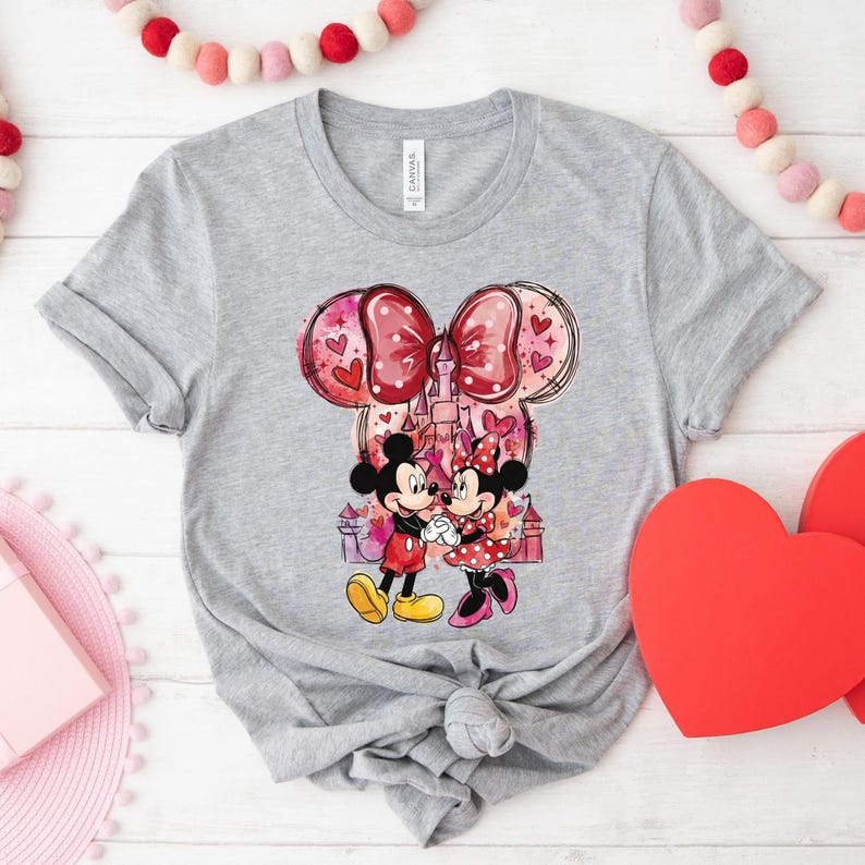 May include: A gray t-shirt featuring a graphic of Mickey and Minnie Mouse holding hands in front of a castle. The design includes a large bow with heart accents and a pink and red color scheme, perfect for Valentine's Day.