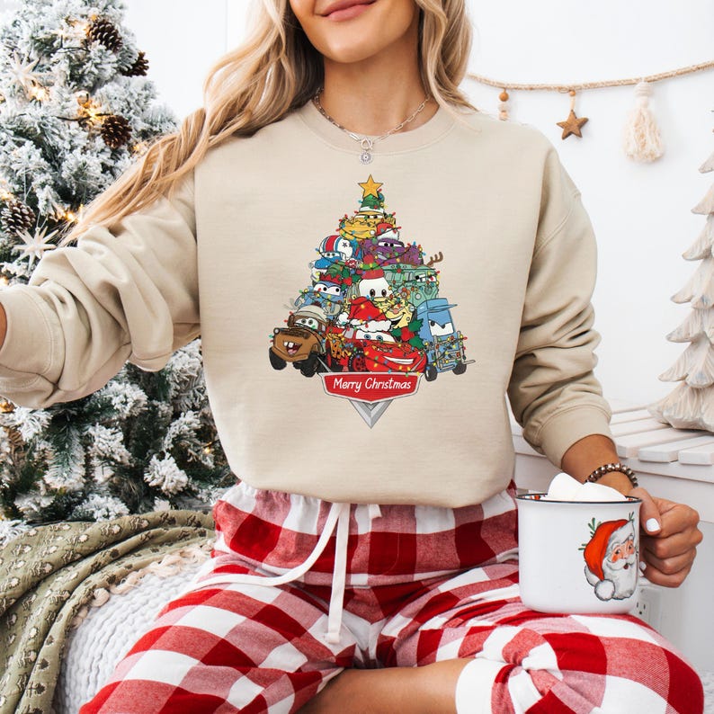 May include: Beige sweatshirt featuring a Christmas tree design made of cartoon cars. The tree is topped with a star and has the words "Merry Christmas" on a banner at the bottom. The person is wearing red and white plaid pajama pants.