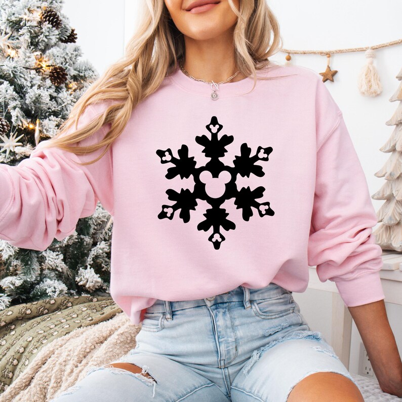 May include: A light pink sweatshirt featuring a black snowflake design with a Mickey Mouse silhouette in the center. The long-sleeved top is made of a soft material, perfect for winter or holiday wear.