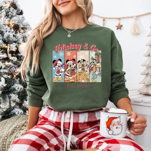 May include: Green sweatshirt featuring Mickey & Co. characters in Christmas attire. The design includes Mickey Mouse, Minnie Mouse, Pluto, and Goofy, with the text "Mickey & Co. -Est. 1928-". The person is wearing red and white plaid pajama pants and holding a Santa mug.