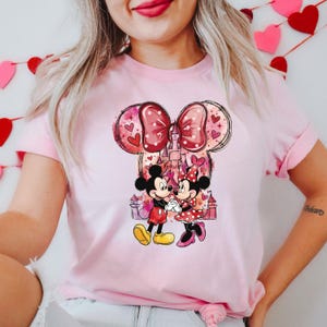 May include: Pink t-shirt featuring a graphic of Mickey and Minnie Mouse holding hands in front of a castle. The design includes a large bow and hearts. The shirt is a light pink color.