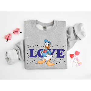 Op de afbeelding: Grijs sweatshirt met een cartoon afbeelding van Donald Duck met een matrozenhoed en het woord "LOVE" in paarse letters. Het sweatshirt is bedekt met kleine zwarte hartjes.