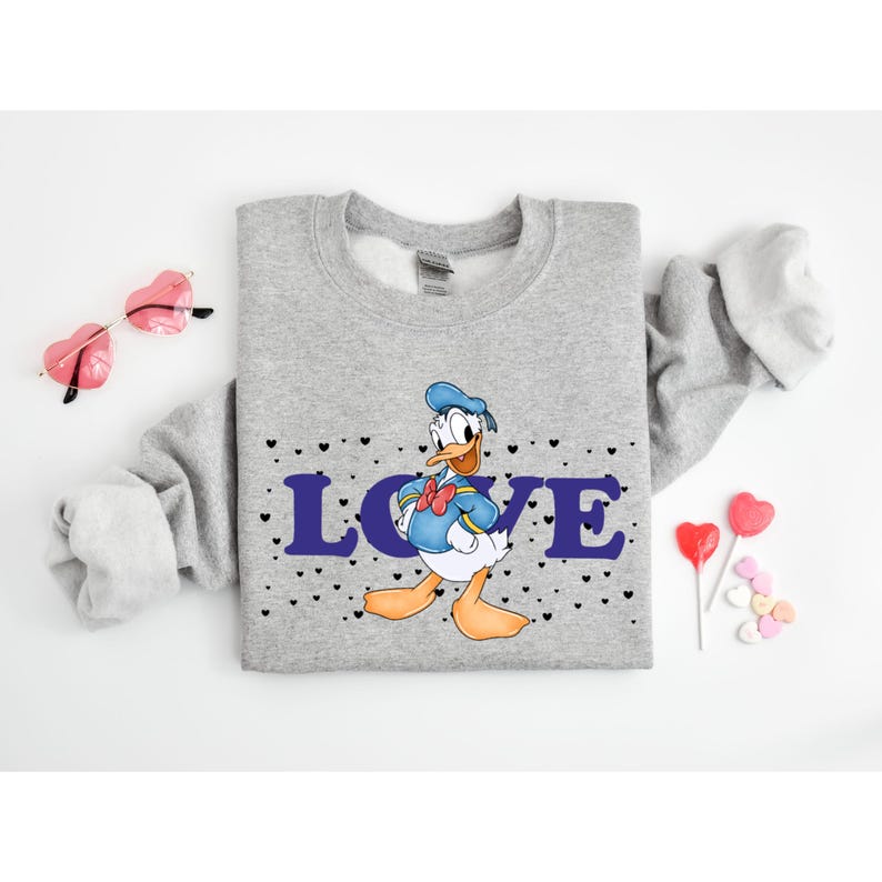 Op de afbeelding: Grijs sweatshirt met een cartoon afbeelding van Donald Duck met een matrozenhoed en het woord "LOVE" in paarse letters. Het sweatshirt is bedekt met kleine zwarte hartjes.