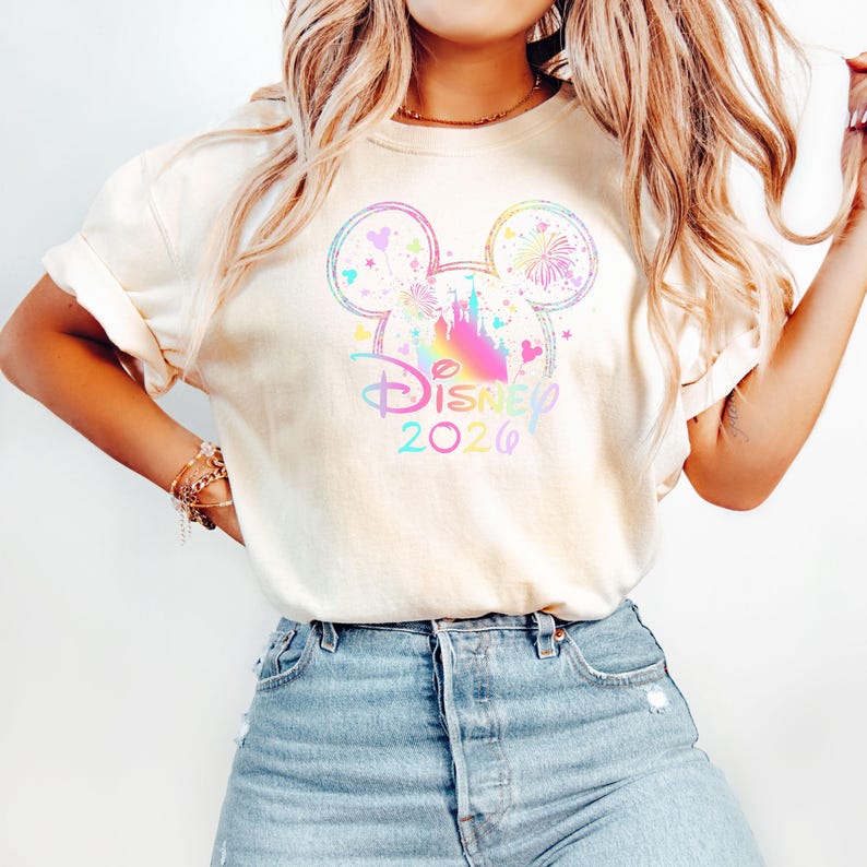 May include: Cream-colored t-shirt featuring a colorful Mickey Mouse head design with a castle, fireworks, and hearts. The text "Disney 2026" is printed below the design in a rainbow gradient. The shirt is paired with blue denim jeans.