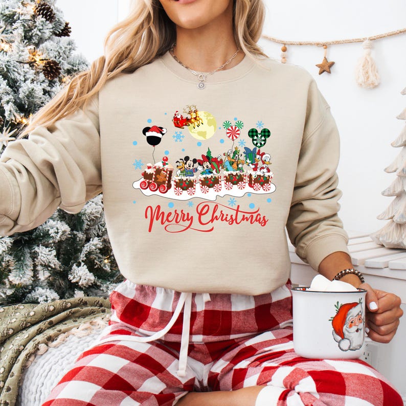 May include: A beige sweatshirt with a Christmas-themed design featuring Mickey Mouse and other Disney characters riding a train. The text "Merry Christmas" is printed below the train.