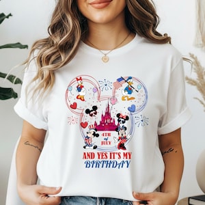 May include: White t-shirt featuring a Mickey Mouse design with patriotic elements. The design includes Mickey and Minnie Mouse, Donald and Daisy Duck, a castle, fireworks, and the text "4th of July AND YES IT'S MY BIRTHDAY".