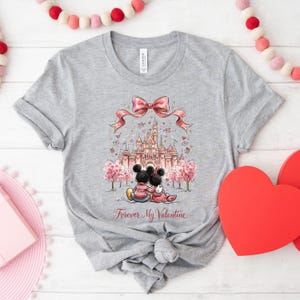 May include: A gray t-shirt featuring a graphic of Mickey and Minnie Mouse in front of a castle, with a pink bow and the text "Forever My Valentine." The design includes pink trees and hearts, perfect for Valentine's Day.