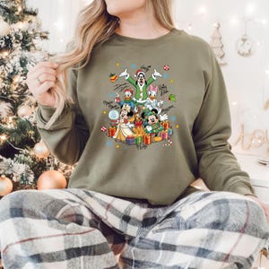 K&ouml;nnte beinhalten: Olivgr&uuml;nes Sweatshirt mit einem festlichen Aufdruck von Disney-Figuren, darunter Mickey, Minnie, Donald und Daisy Duck, umgeben von Geschenken und Schneeflocken. Das Design enth&auml;lt Charakterunterschriften und Weihnachtselemente.