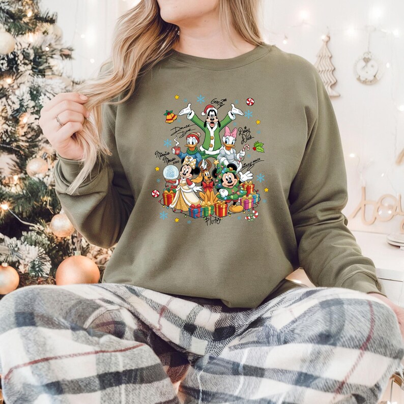 K&ouml;nnte beinhalten: Olivgr&uuml;nes Sweatshirt mit einem festlichen Aufdruck von Disney-Figuren, darunter Mickey, Minnie, Donald und Daisy Duck, umgeben von Geschenken und Schneeflocken. Das Design enth&auml;lt Charakterunterschriften und Weihnachtselemente.